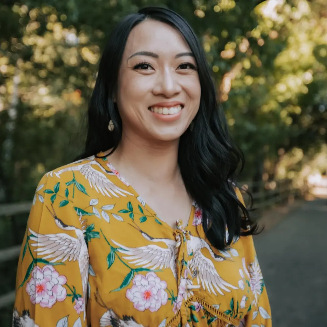 A woman in yellow and white floral shirt standing next to trees.